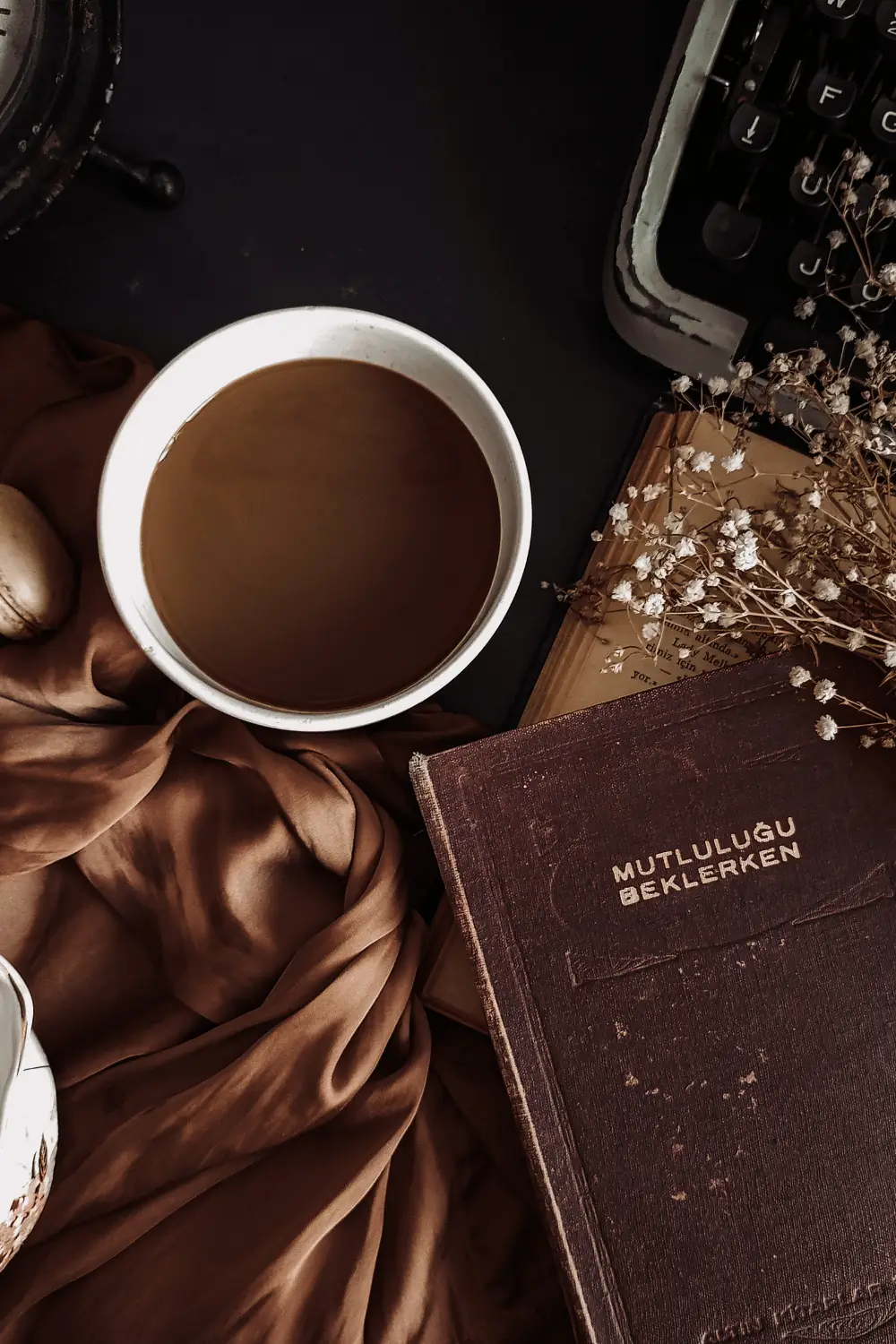 A warm, moody flat lay featuring a cup of coffee beside an old brown book, a vintage typewriter, dried flowers, and soft brown fabric—creating a cozy, poetic atmosphere perfect for writing or reflection.