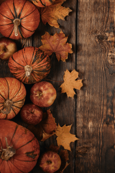 Overhead view of rustic autumn scene with orange pumpkins, red apples, and dried maple leaves arranged on a dark wooden table.