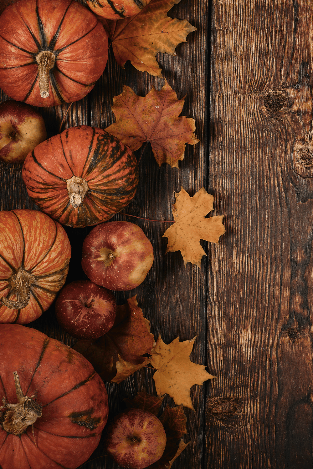 Overhead view of rustic autumn scene with orange pumpkins, red apples, and dried maple leaves arranged on a dark wooden table.