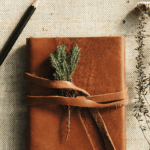 Brown leather journal wrapped with a strap and decorated with a small sprig of greenery, lying on a textured fabric surface beside a pencil and dried wildflowers.
