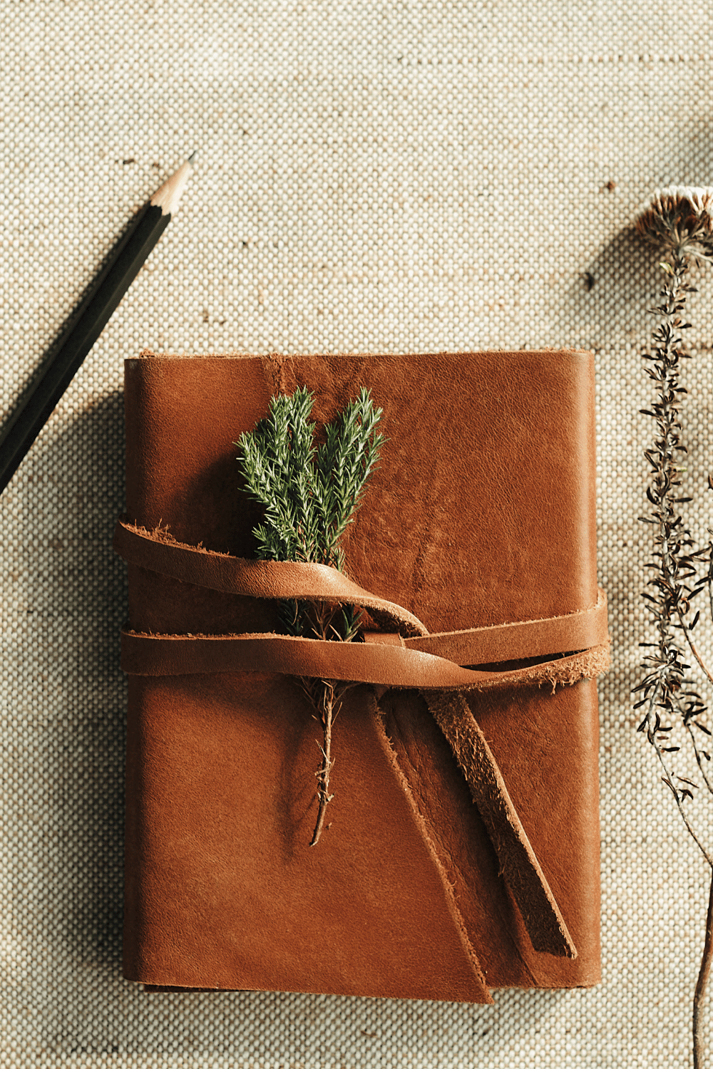 Brown leather journal wrapped with a strap and decorated with a small sprig of greenery, lying on a textured fabric surface beside a pencil and dried wildflowers.
